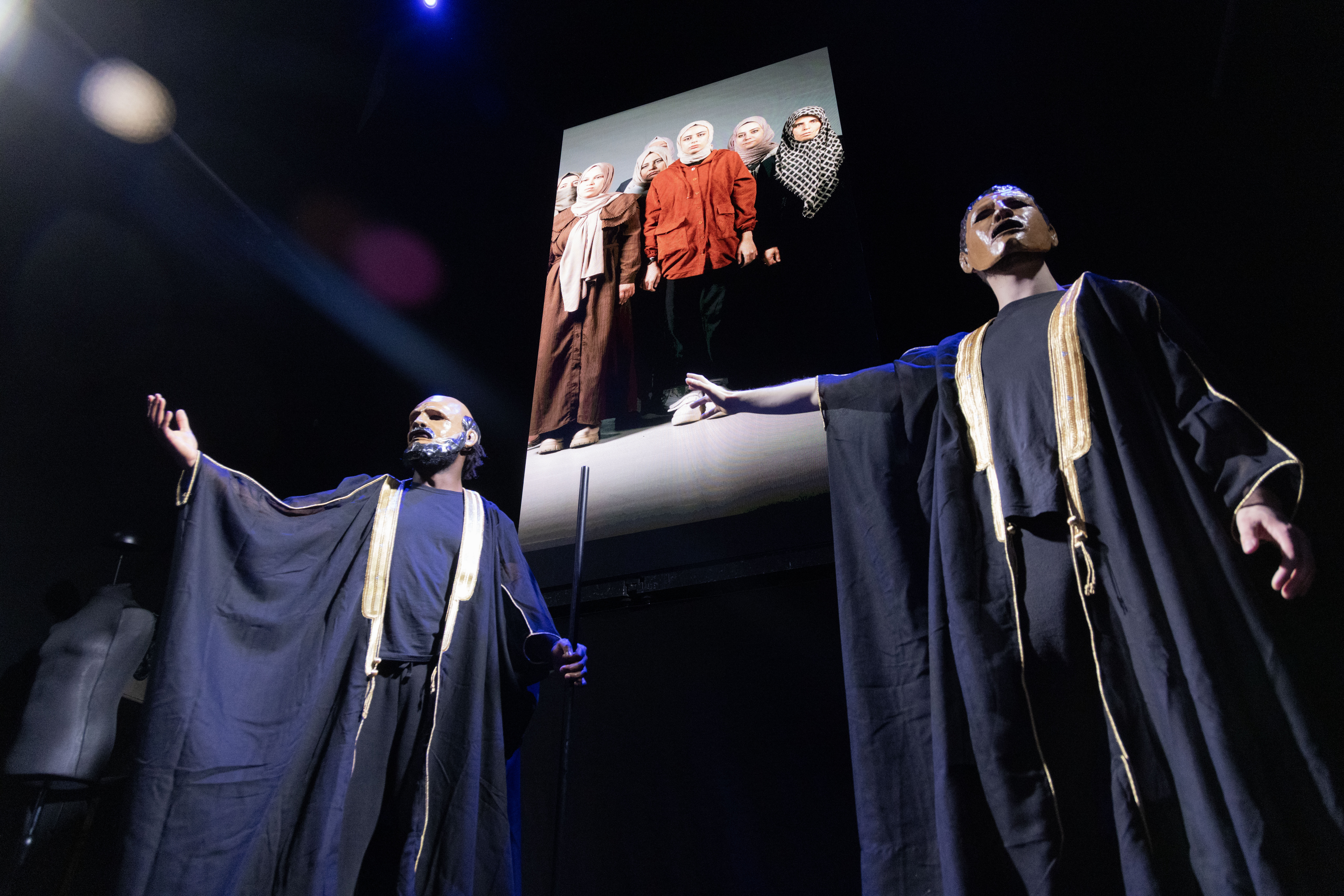 Two masked men in front of a chorus of women on a screen